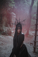 Young woman in witch stands in black dress and crown on her head in dark and foggy forest.