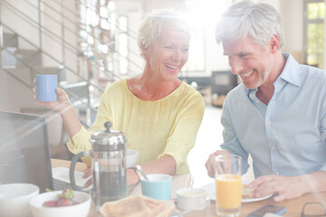 Older couple laughing together at breakfast table with laptop