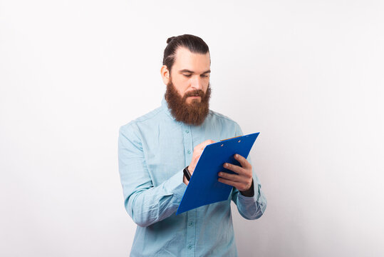 Serious Bearded Man Is Writing Something On A Paper Holder Board While Standing Over White Background.