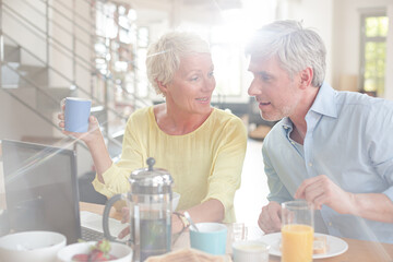 Older couple laughing together at breakfast table with laptop