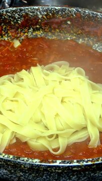 Spaghetti put in savory tomato sauce with vegetables, simmer in frying pan on gas stove. Recipe for making pasta or tagliatelle in home kitchen. Vertical video. Close-up.