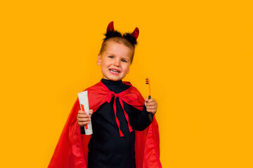 One little toddler boy in a carnival costume with toothpaste and an orange brush for Halloween is isolated on a yellow background. Medicine, dental hygiene, holidays concept.