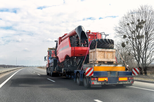 Heavy Industrial Truck With Semi Trailer Platform Transport Disassembled Combine Harvester Machine On Common Public Highway Road On Sunset Sunrise Day. Agricultural Equipment Transportation Service