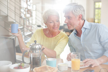 Older couple laughing together at breakfast table with laptop