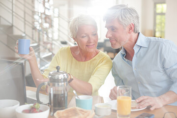 Older couple laughing together at breakfast table with laptop