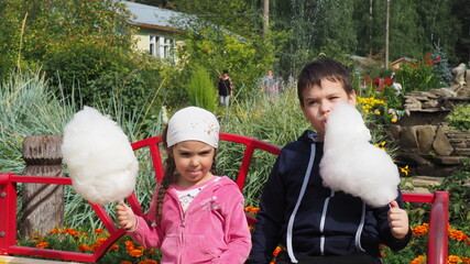 Happy cute boy and girl eating cotton candy floss in the park near the fountain sitting on a bench....