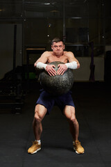 handsome muscled young man with a naked torso in a gym holding a huge stone ball with great effort