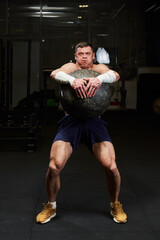 Muscled young man with a naked torso in the gym holding a huge stone ball with huge effort