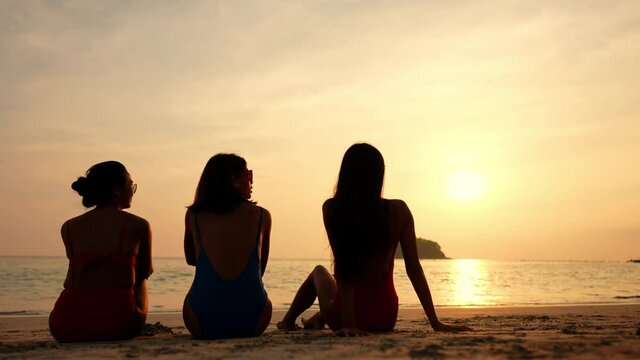 Group of Asian woman friendship in colorful swimwear sitting on the beach and talking together at summer sunset. Female girl friends enjoy and having fun outdoor activity lifestyle on summer vacation