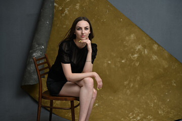 A full-length photo of a young brunette girl sits on a chair in the studio on a gray-golden background