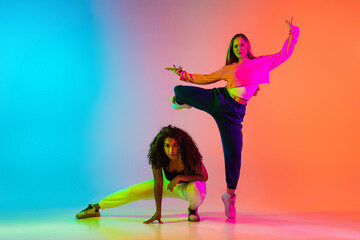 Two beautiful girls dancing hip-hop on colorful gradient background in neon lights