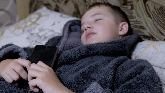 Smiling Child in Home Dressing Gown falls asleep with Smart Phone in Hands. Happy boy kissing mobile phone screen before bedtime. A teenager is dependent on modern gadgets and tablets. 4K. Close up.