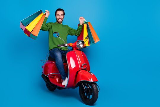Full size photo of excited guy wear green sweater ride moped raise fists in victory after shopping isolated on blue color background