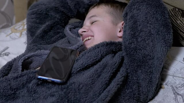 Yawning Child in Home Gray Dressing Gown Falls asleep with Smartphone in Hands. A smiling boy holds a mobile phone in his hands. A teenager is dependent on modern gadgets and tablets. 4K. Close up.