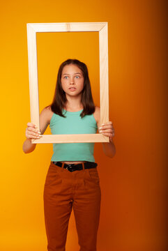 a cute teenage girl of 15 years old shows a smile and sadness through a wooden frame holding her in her hands on a yellow background. Knowledge Day