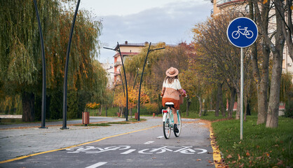 Young woman with curly hair in hat and stylish dressed riding bicycle on bike lane in urban park or...