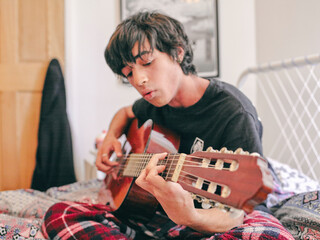 Teenage boy playing acoustic guitar on bed
