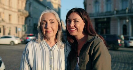 So similar. Waist up portrait view of the happy mother and daughter bonding and embracing with each other while standing at the sunny street and smiling to the camera