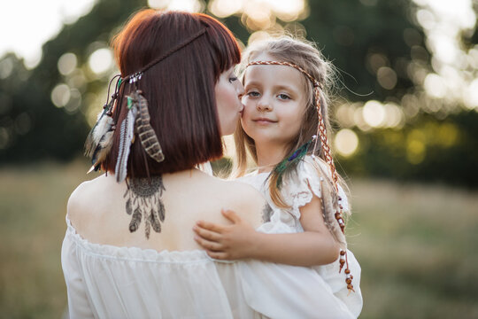 Rear view of lovely caring woman mother, holding on hands her pretty cute daughter and enjoying summertime. Boho woman with dream catcher tattoo on back, hugging and kissing her little child in field