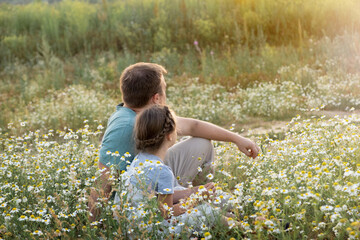 a father and a little daughter sit side by side on the grass in a chamomile field on a sunny summer day and look into the distance at the sunset. Family happiness