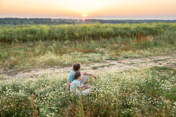 a father and a little daughter sit side by side on the grass in a chamomile field on a sunny summer day and look into the distance at the sunset. General plan