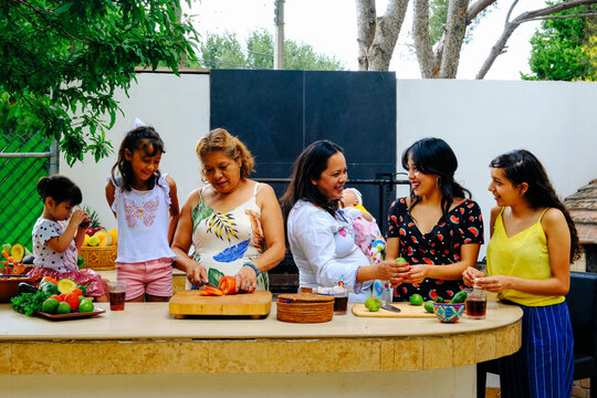 Multi-generational smiling women and girls preparing salsa together at kitchen counter in backyard during weekend