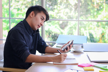 Busy businessman in black shirt working on office desk, taking notes while talking with customer on mobile phone, and the other hand holding a calculator, writing information data on paper.
