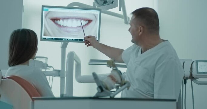 A female patient is sitting in the dentist's chair and looking at the monitor screen with a human mouth with teeth while a male dentist explaining something and holding a pointer to show teeth.