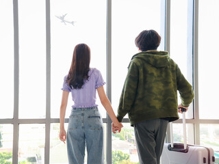 Asian young tourist lover couple hold each other hand, looking through window, standing with luggage suitcase from behind, waiting airline flight at airport terminal, romantic trip on vacation.