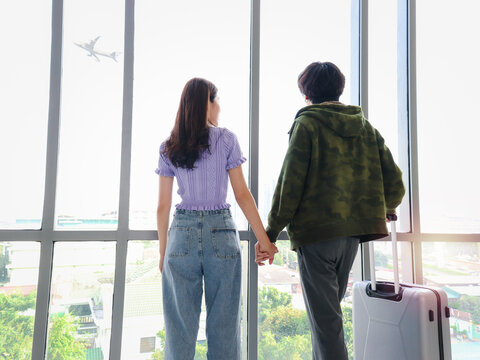 Asian Young Tourist Lover Couple Hold Each Other Hand, Looking Through Window, Standing With Luggage Suitcase From Behind, Waiting Airline Flight At Airport Terminal, Romantic Trip On Vacation.