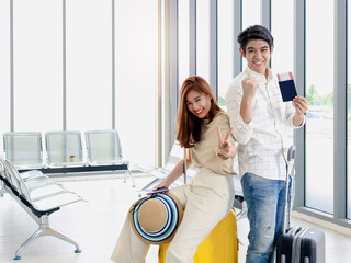 Obraz premium Asian young traveler lover couple holding passport and boarding pass (mockup), standing with luggage suitcase, waiting airline flight at airport terminal, have fun together, romantic trip on vacation.