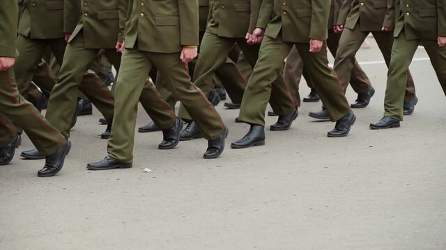 Military officers are marching in straight ranks across square. Close-up of legs in green pants with matching black shoes.