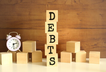 Five wooden cubes stacked vertically to form the word DEBTS on a brown background.