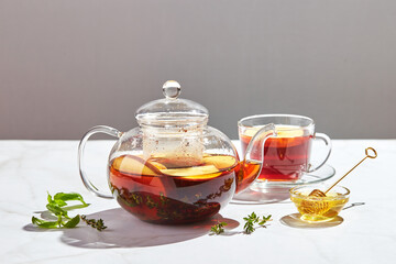 Fruit tea with apples and thyme and honey in glass teapot and cup on white background with hard shadows