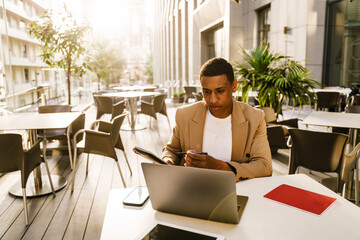 Young black man writing down notes while working with laptop at cafe