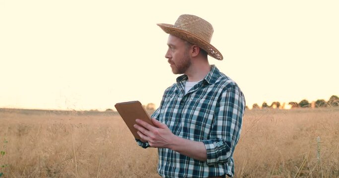 Waist up of the caucasian adult man in the plaid shirt and hat looking at the camera and thinking while taping on the tablet device. Portrait of farmer concept