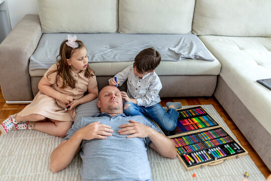 Adorable kids having fun with a sleeping father at home. Kids are having fun drawing on their father's face while he his sleeping on the floor. April fools prank. Lifestyle.