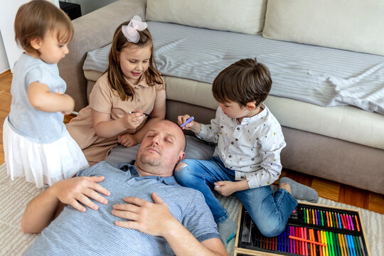 Children carefully paints their father's face during family games together. Siblings painting their dad face with a brush, watercolour and marker while he sleeps on the floor at home. Family bonding.