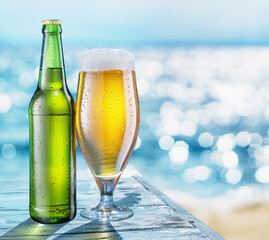 Bottle of beer and beer glass with condensation on the wooden table. Blurred sparkling sea at the background. Summer vibes.
