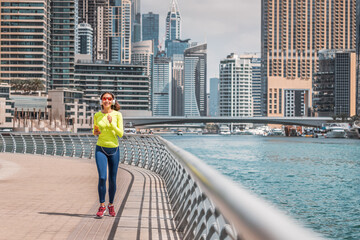 Happy woman wearing headphones and sunglasses trains and does running and fitness on the sidewalk of the embankment in the Dubai Marina area