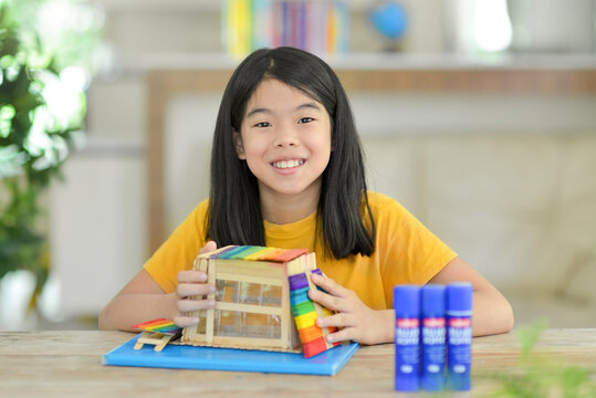 Asian child doing popsicle stick craft.School kid with colorful homemade wooden sticks house.Creative idea for fun activity in class.Colorful sticks.Glue on table.Homeschooling projects.