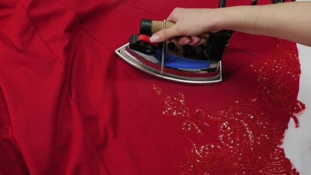 A young woman dressmaker irons a red openwork dress in a sewing workshop, sewing and making new clothes.