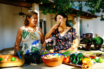 Happy mature woman looking at young daughter eating tortilla chips with Mexican salsa in backyard on sunny weekend - obrazy, fototapety, plakaty