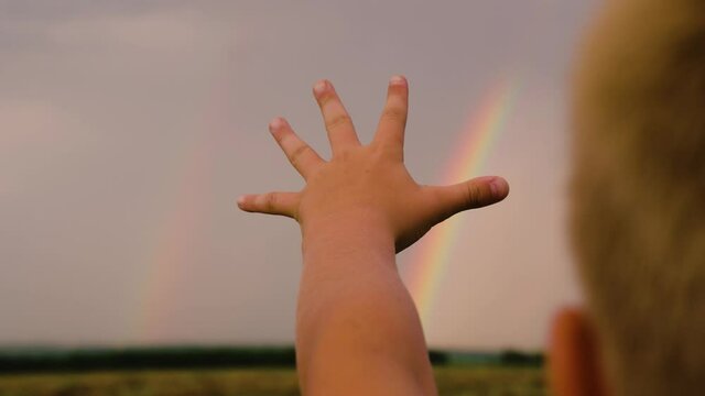 The child dreams of touching a multi-colored rainbow in the sky. Happy family concept. Happy child, boy stretches out his hand to the rainbow against the blue sky. Family walks in the park after rain.