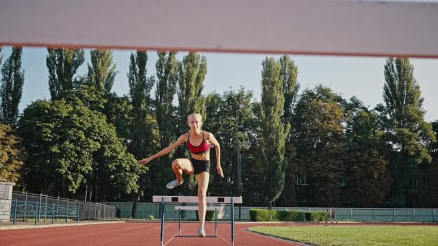 Professional athlete runs a sprint with obstacles, different running techniques. Morning workout in the sunshine at the city stadium. The blonde girl trains in the summer at the open stadium