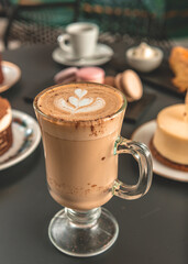 Capuchino con arte latte en un primer plano acompañado de distintos tipos de tortas con foco diferenciado.