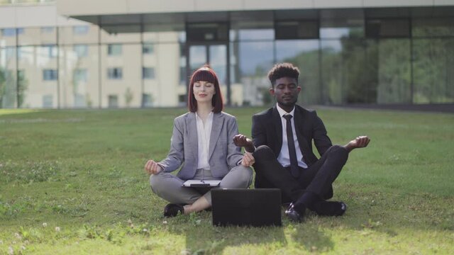 Office yoga, staff recreation and mindfullness concept. Two young mutli-cultural employees in a lotus pose are sitting on green grass and meditate with eyes closed outside modern office behind.