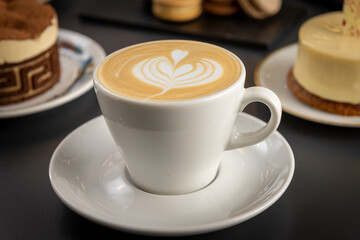 Cafe con arte latte en un primer plano acompañado de distintos tipos de tortas con foco diferenciado.