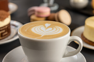Café  con arte latte en un primer plano acompañado de distintos tipos de tortas con foco diferenciado.