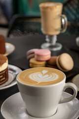 Café  con arte latte en un primer plano acompañado de distintos tipos de tortas con foco diferenciado.